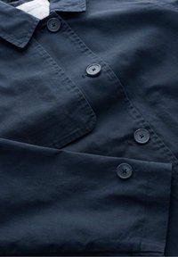 Granatowa koszula zapinana na guziki z widocznym kołnierzykiem, przednią kieszonką oraz czarnymi guzikami z przodu i przy mankietach.