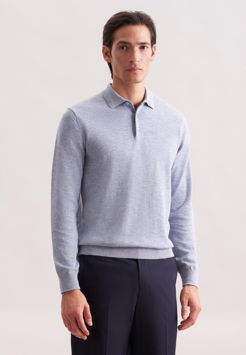 Mann in einem hellgrauen Langarm-Polo-Pullover und dunklen Navy-Hosen, der vor einem schlichten hellen Hintergrund steht.
