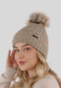 Bonnet tricoté de couleur fauve avec une texture côtelée, doté d'un pompon en fausse fourrure sur le dessus et d'une petite étiquette noire sur le côté.