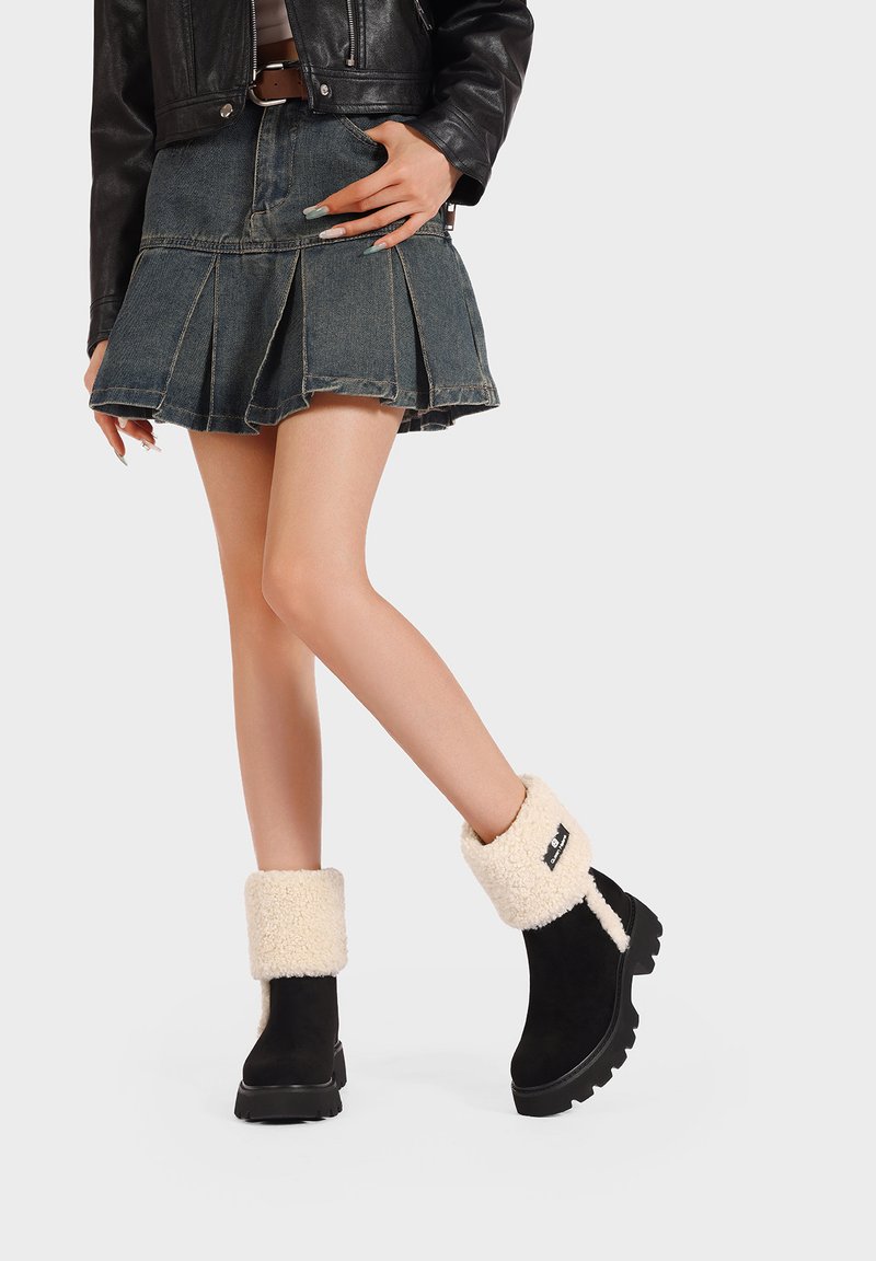 Stivaletti neri in suede con risvolto in pelliccia bianca, caratterizzati da una suola spessa e un'etichetta con logo laterale, abbinati a una gonna plissettata in denim.