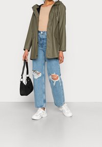 Person, der einen olivgrünen Regenmantel, ein beiges geripptes Oberteil, hellblaue, zerrissene Jeans und weiße Sneaker trägt und eine schwarze Handtasche hält.