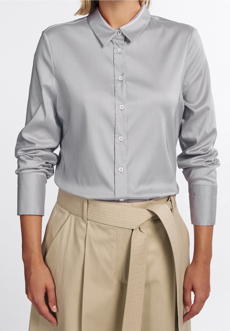 Femme portant une chemise à manches longues gris clair boutonnée, rentrée dans un pantalon beige taille haute avec une large ceinture en tissu nouée à la taille.