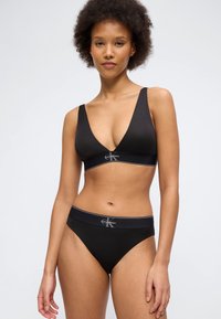 Γυναίκα με κοντά σγουρά μ αλλιά που φοράει μαύρο bralette Calvin Klein και ταιριαστό εσώρουχο, στέκεται μπροστά σε μια απλή λευκή φόντο.