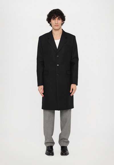 Schwarzer langer Mantel mit taillierter Passform, mit Revers, drei Knöpfen und seitlichen Taschen, kombiniert mit grauen Karohosen und schwarzen Schuhen.