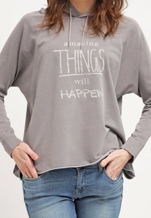 Personne portant un sweat à capuche gris avec le texte "amazing THINGS will HAPPEN" et un jean bleu, les mains reposant dans les poches avant.