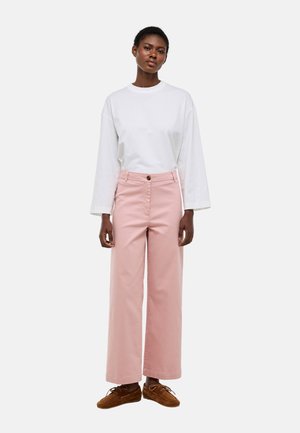 Femme debout portant un haut blanc à manches longues, un pantalon large taille haute rose et des chaussures montantes marron, sur un fond uni.