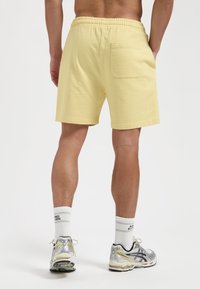Lichtgele katoen shorts met een elastische tailleband, met een enkele achterzak. Draag met witte sokken en grijze sport schoenen.