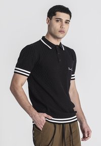 Černé polo tričko s texturou, bíle pruhovaný límec a manžety, s kapsou a vyšitým detailem na hrudi.