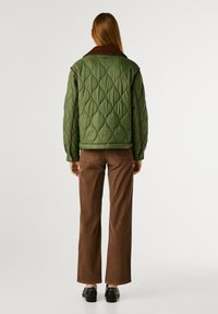 Giacca trapuntata verde con colletto marrone a contrasto, abbinata a pantaloni in corduroy marroni. Presenta tasche laterali e un motivo testurizzato.