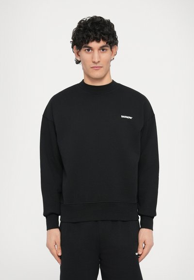 Melns džemperis no mīksta auduma ar apaļu kakla izgriezumu, garām piedurknēm un nelielu baltu logo kreisajā krūtīs. Vienkārša, brīvā piegriezuma.