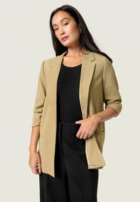 Beige szabott zakó, kihajtott gallérral és felgöngyölt ujjakkal, fekete bordás felsővel és fekete, magas derekú nadrággal párosítva.