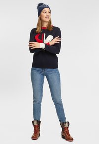 Granatowy sweter z czerwonym i białym wzorem górskim, zestawiony z niebieskimi dżinsami i brązowymi butami z futrzanymi akcentami. Model nosi niebieską wełnianą czapkę.