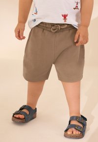 Hellbraune Shorts mit elastischem Bund und Kordelzug, kombiniert mit blauen Sandalen mit doppelten Schnallenriemen und Korksohle.