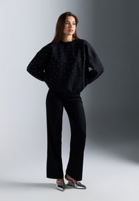 Sudadera negra con adornos dispersos, combinada con pantalones negros de pierna ancha y zapatos plateados de punta afilada, contra un fondo degradado.