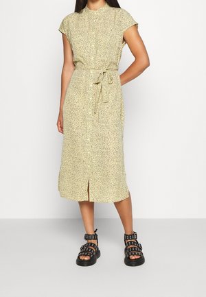 Žluté šaty s květinovým vzorem, krátkými rukávy, páskem v pase a zapínáním na knoflíky vpředu; doplněné o černé sandály s pásky.