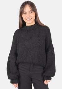 Schwarzer Strickpullover mit hohem Kragen, weiten Ärmeln und strukturiertem Muster. Kombiniert mit schwarzer Hose.