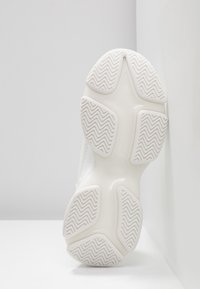 Semelle d'une basket blanche avec des coussinets de grip texturés et un motif de bande de roulement en zigzag, montrée sur une surface blanche angulaire.