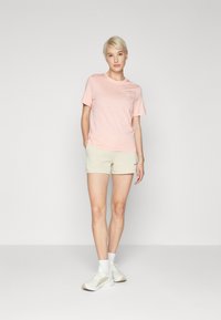 Svijetlopinkana pamučna majica s kratkim rukavima i okruglim izrezom, u kombinaciji s bež šortsevima. Model stoji na jednobojnoj pozadini, odjeven u bijele tenisice.