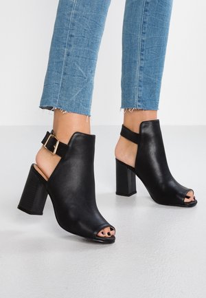 Czarne botki za kostkę z odkrytymi palcami, wykonane z gładkiej skóry, z blokowym obcasem oraz regulowanym paskiem na kostkę z akcentem w postaci złotej klamry.