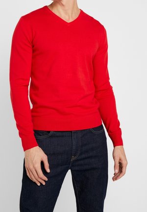 Pull rouge à manches longues et col en V en tissu tricoté lisse, avec des poignets et un ourlet à côtes. Porté avec un jeans bleu foncé.