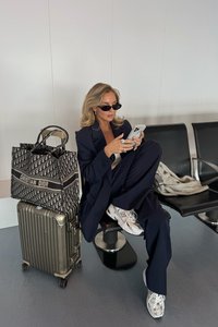 Schwarze und beige Christian Dior Tote-Tasche neben einem silbernen Hartschalenkoffer mit Rollen. Person in marineblauem Anzug und New Balance Sneakers schaut auf ihr Handy.