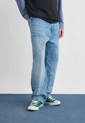 Asmuo dėvi šviesiai mėlynas džinsines kelnes su siuvinėjimu, žalias drobines sportbačius su baltais raišteliais, juodą marškinėlį ir mėlynus languotus ilgomis rankovėmis marškinius.