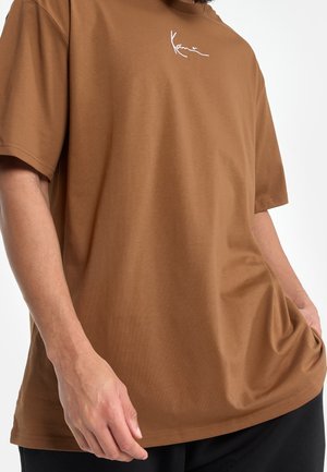 Camiseta de algodón marrón con un corte holgado, mangas cortas y un logo de firma blanco en el pecho. Textura suave con un diseño minimalista.