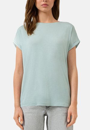 Woman wearing a light blue short-sleeve T-shirt and light gray jeans standing against a plain background.