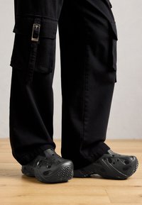 Czarne spodnie cargo z bocznymi kieszeniami, w połączeniu z czarnymi klapkami typu slip-on z perforowanym wzorem, noszone na drewnianej podłodze.