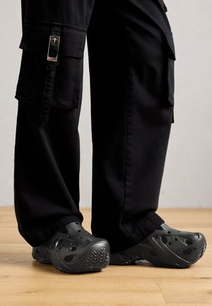 Black cargo pants with side pockets paired with black slip-on clogs featuring perforated design, worn on wooden floor.