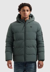 Man in een donkergroene gewatteerde jas met capuchon en ritssluiting aan de voorkant, rechtopstaand tegen een vlakke lichte achtergrond.