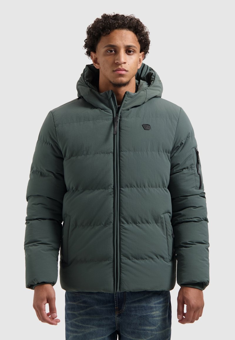 Man in een donkergroene gewatteerde jas met capuchon en ritssluiting aan de voorkant, rechtopstaand tegen een vlakke lichte achtergrond.