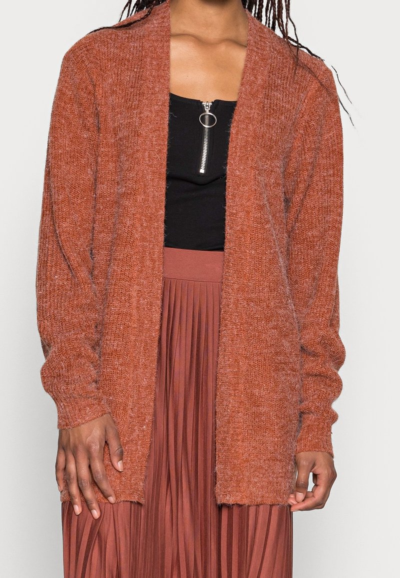 Cardigan a maglia rosso mattone con apertura sul davanti, maniche lunghe e una finitura morbida e texturizzata, abbinato a una maglietta nera e a una gonna marrone plissettata.