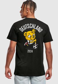 Juodas medvilninis marškinėliai su geltonu animaciniu lokiu, žaidžiančiu futbolą, viršuje užrašas "DEUTSCHLAND 2024" ir žaismingi grafikos detalės.