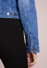 Detailní záběr osoby v modré džínové bundě a černých kalhotách, jejíž ruka spočívá vedle těla na neutrálním pozadí.