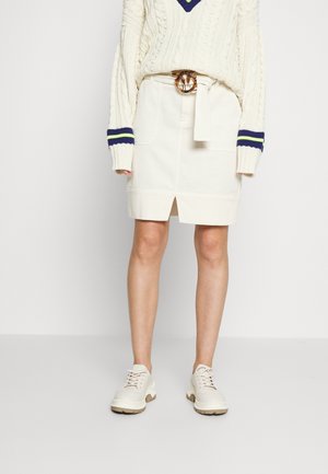 Personne portant une jupe blanche avec une fente devant, un pull en maille torsadée crème avec des rayures bleu marine et citron vert, et des chaussures blanches à lacets sur un fond clair.