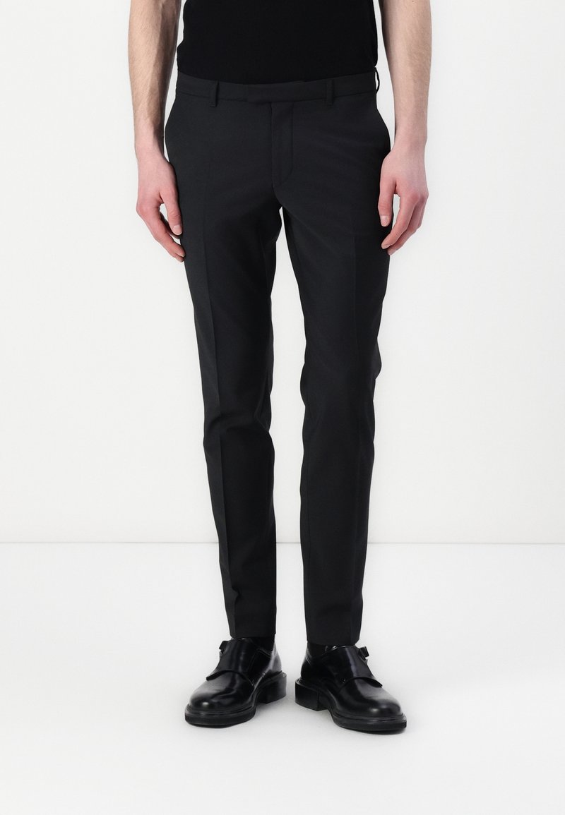 Svarta skräddarsydda byxor med smal passform, slät front, jämn textur och minimala detaljer, tillsammans med svarta skor, stående mot en vit bakgrund.