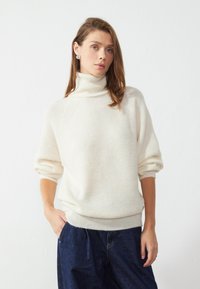 Pull en maille côtelée crème à col roulé, avec des épaules tombantes, des manches mi-longues et une coupe ajustée. Associé à un jean large bleu foncé.
