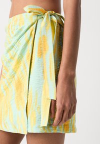 Falda de estilo envolvente en tejido ligero con una base verde menta y patrones de rayas amarillas y naranjas; incluye una cinturilla atada.