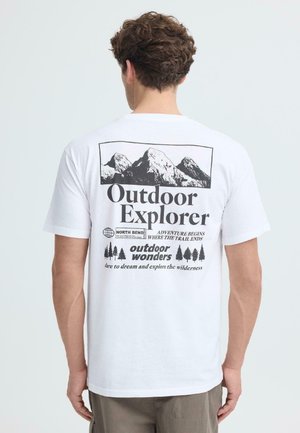 Man draagt een wit T-shirt met zwarte grafische afbeeldingen van een berg en een boom, met de tekst "Outdoor Explorer, North Bend, outdoor wonders" op de achterkant.