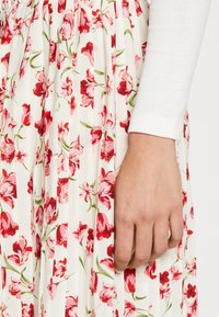 Blommig plisserad kjol med vit bas och mönster av röda, rosa och gröna blommor; har en veckad midja och mjukt, texturerat tyg.