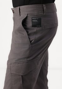 Pantalones cargo grises hechos de una tela duradera, con bolsillos laterales, acentos de costura y una etiqueta de la marca en la cinturilla.