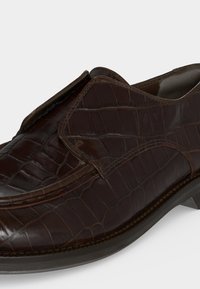 Mocassin en cuir marron foncé avec motif crocodile, surpiqûres moc toe et design slip-on sur un fond clair uni.