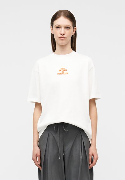 Νέα γυναίκα που φοράει λευκό φαρδύ T-shirt με πορτοκαλί κείμενο "IT'S BETTER IN STIEGLITZ" και φαρδιά γκρι πλισέ παντελόνια, σε απλό φόντο.