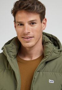 Oliwkowa kurtka puchowa z kapturem, z zamkiem błyskawicznym i małym białym logo na piersi. Noszona na brązowej koszuli.