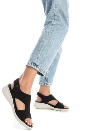 Sandalias negras de punto con punta y talón descubiertos, con una suela blanca. Combinadas con unos jeans azul claro arremangados, que muestran detalles recortados.