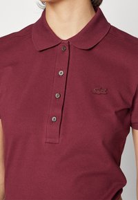 Polo shirt bordeaux en tissu texturé, manches courtes et col classique. Comprend trois boutons et un petit logo brodé.