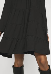 Robe noire à volants avec une texture fluide, comprenant des coutures froncées et une coupe mi-longue, associée à des bottes noires montantes jusqu'au genou.