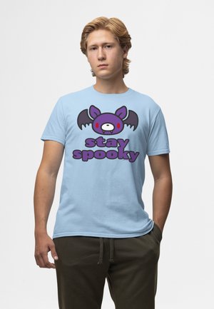 Jeune homme portant un t-shirt bleu clair avec une chauve-souris dessin animé violette et le texte « stay spooky », associé à un pantalon foncé, debout devant un fond uni.