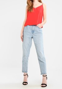 Haut rouge sans manches à col en V, associé à un jean taille haute bleu clair. Sandales noires à bride à la cheville complètent la tenue.